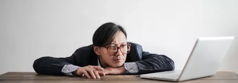 Man in glasses staring thoughtfully at a laptop on a wooden table.